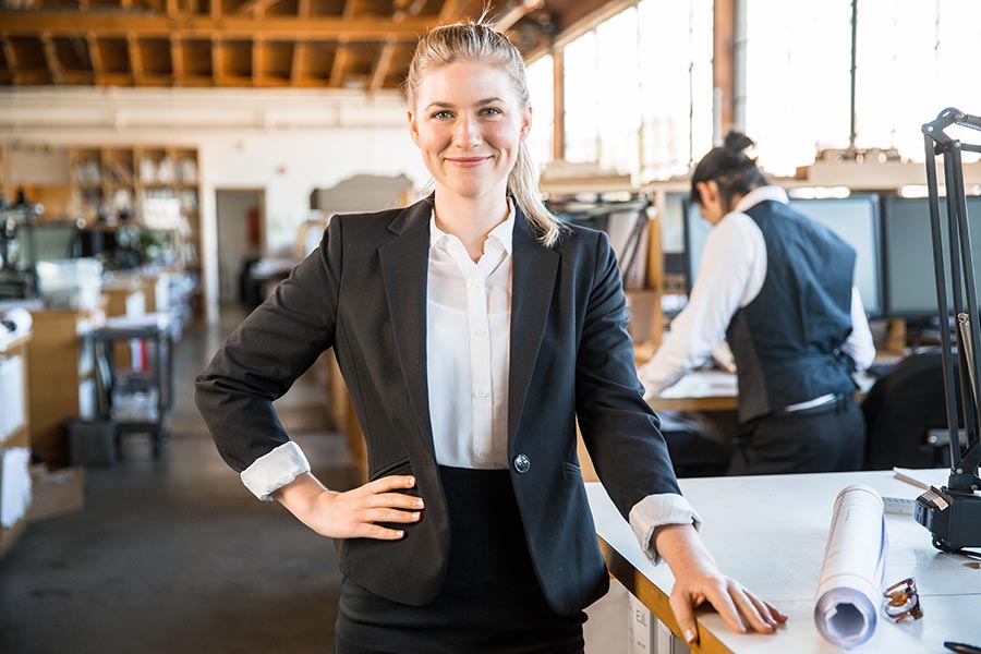 Business Insurance - Business Owner in a Garment Workshop Stands at a Worktable Wearing a Black Suit, Smiling